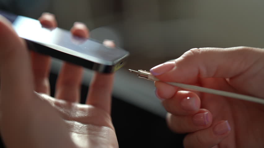 Close-up of female hands plugging white lightning cable into smartphone to charge battery, unplugging, showing connecting and disconnecting device for power and data transfer. Shooting in slow motion.