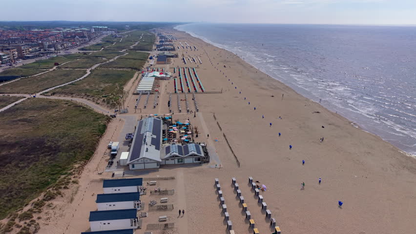 A magnificent wide-angle aerial photograph capturing a high-altitude drone overview of the vibrant coastal resort of Zandvoort. The sweeping high-angle composition reveals a long sandy beach stretching along the shimmering North Sea coast, featuring neatly arranged rows of colorful beach cabins, beach houses, pavilions, and outdoor dining terraces equipped with solar panels. Parallel to the shore, grassy coastal dunes and paved paths separate the beach infrastructure from the adjacent coastal city skyline in the distance. Bright afternoon sunlight reflects off the gentle sea waves, illuminating the wide sandy coast and green dune landscape under a clear, hazy blue summer sky. This panoramic perspective beautifully documents European coastal geography, resort urban planning, dune conservation areas, and popular Dutch seaside tourism destinations.