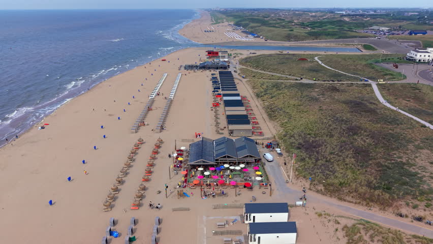 A magnificent wide-angle aerial photograph capturing a high-altitude drone overview of a coastal beach resort near Zandvoort. The sweeping composition showcases a lively beach pavilion with colorful patio umbrellas, surrounded by neatly arranged rows of beach cabins and thatched straw parasols along the wide sandy shoreline. On the left, gentle North Sea waves break against the sandy beach, while on the right, expansive grassy sand dunes stretch toward inland walking paths and parking areas. In the distant background, a small coastal water channel cuts through the dunes under a clear blue summer sky. Bright afternoon daylight illuminates the vibrant seaside scenery, highlighting rich textures of sand, sea, and coastal vegetation. This high-angle perspective beautifully documents European coastal geography, resort architecture, dune landscape conservation, and popular Dutch seaside tourism.
