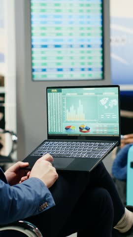 Vertical composition: Businessman reviewing company reports on laptop in airport lounge during business trip, analyzing data, KPIs and budget performance.