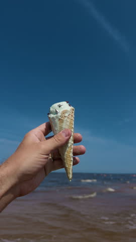 Hand holding melting ice cream cone against bright blue sky with beach and child in soft focus