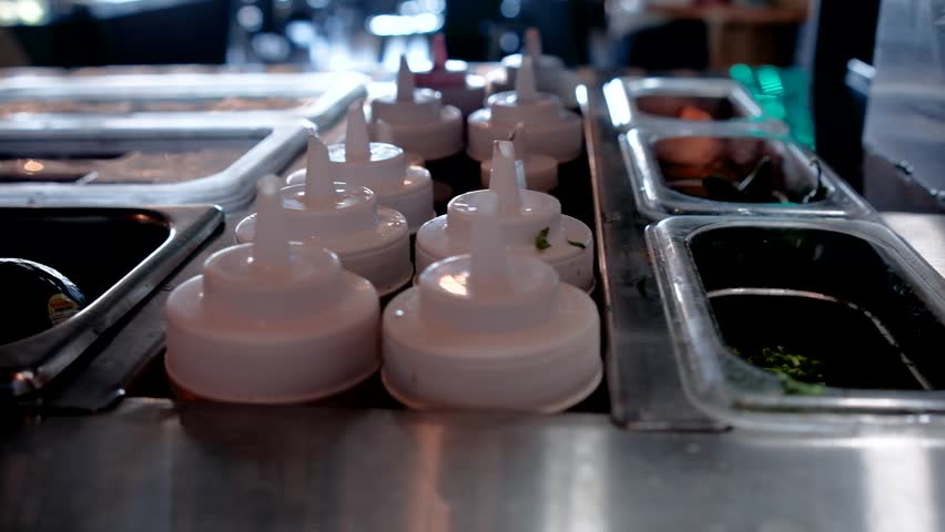 Shallow focus view along a stainless steel restaurant prep counter with rows of squeeze bottles metal ingredient bins sauces and toppings arranged for Mexican seafood service