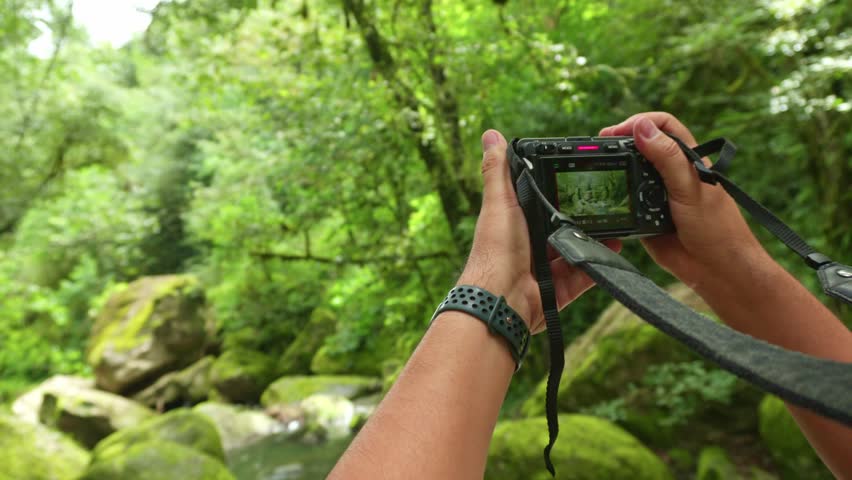 Over the shoulder view of a man using a digital camera to capture a lush forest river landscape