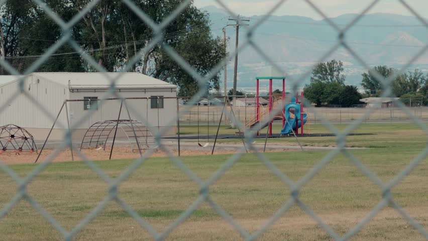 Steady handheld shot looking through a chain link fence toward a playground and relocatable building.