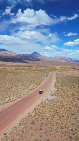 Tracking shot following a lone vehicle driving along a remote dirt road through a vast high altitude desert plateau with volcanic peaks rising under a dramatic blue sky