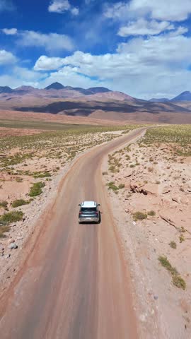 Aerial tracking shot following a lone car driving along a remote desert road through a vast arid plateau with layered Andean mountain ranges under a dramatic blue sky