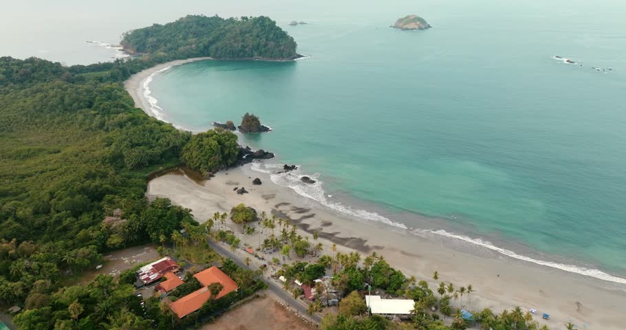 Tropical beach and forested Quepos coastline beside the Pacific Ocean in Puntarenas, Costa Rica, with sandy shore, rocky islets and turquoise water, aerial dolly backwards.