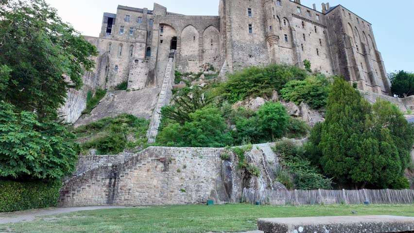 Lateral view of Mont Saint-Michel Abbey towering above steep fortified walls and lush greenery in Normandy, France.
