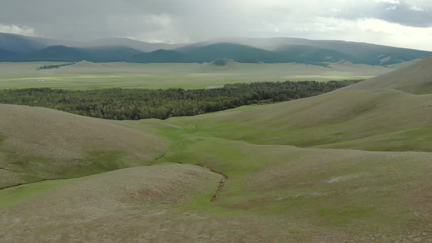 Aerial drone view shows Mongolian forest meadow and winding stream across green plains. Boreal woodland meets grassland creek corridors within pristine remote wilderness. The overhead perspective highlights the transition between woodland, open pasture, and the sinuous freshwater channel. This remote landscape represents a natural meeting point of forest and steppe ecosystems in Mongolia. The scene supports concepts of biodiversity, watershed ecology, habitat corridors, conservation, and sustainable land use. Green vegetation and flowing water provide versatile imagery for geography, environmental science, travel, and wilderness projects.