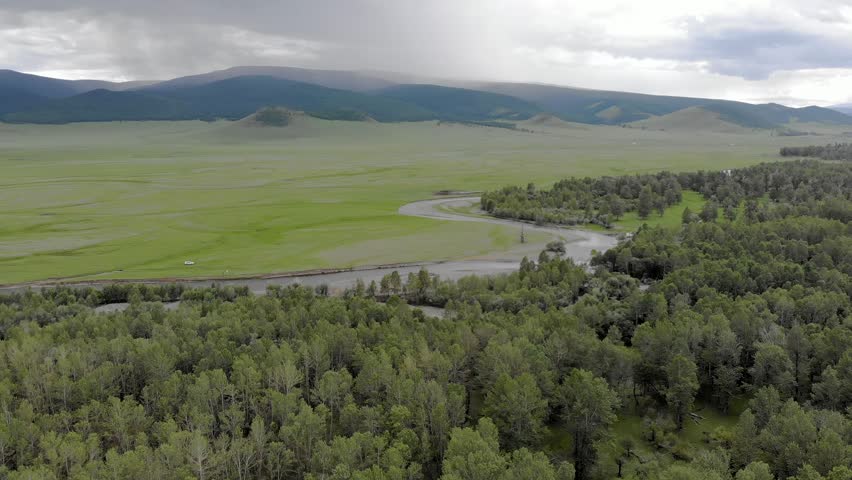 Aerial drone view shows Mongolian forest meadow and winding stream across green plains. Boreal woodland meets grassland creek corridors within pristine remote wilderness. The overhead perspective highlights the transition between woodland, open pasture, and the sinuous freshwater channel. This remote landscape represents a natural meeting point of forest and steppe ecosystems in Mongolia. The scene supports concepts of biodiversity, watershed ecology, habitat corridors, conservation, and sustainable land use. Green vegetation and flowing water provide versatile imagery for geography, environmental science, travel, and wilderness projects.