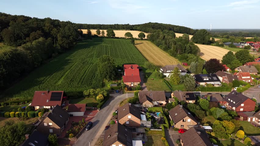 Sunset light bathes German suburb bordering green cornfield and golden wheat fields, red-tiled houses with solar panels line quiet tree-shaded street below rolling hills. Aerial drone flyover shot