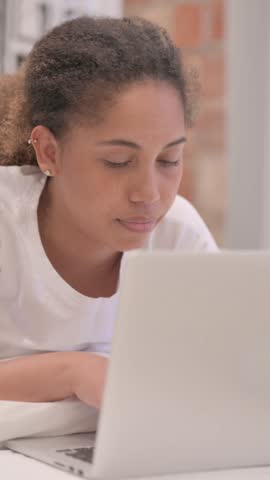 African American Woman Smiling at Camera while Working on Laptop while Lying on Stomach in Bed, vertical video