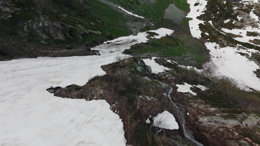 Aerial footage captures the transition from snow-covered terrain to lush green valleys, showcasing a serene mountain landscape with a reflective pond