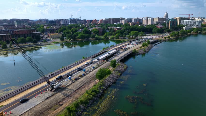 Aerial drone shot flying over a lake causeway road and railway under construction with a crane, traffic, and the Madison, Wisconsin city skyline in the background on a sunny day.