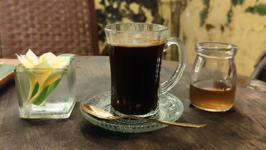 Serving signature Robusta black coffee from Java, Indonesia. a shot showing sugar syrup being added to a glass of coffee. The setting is a quiet, warm room perfect for relaxing and enjoying the coffee.