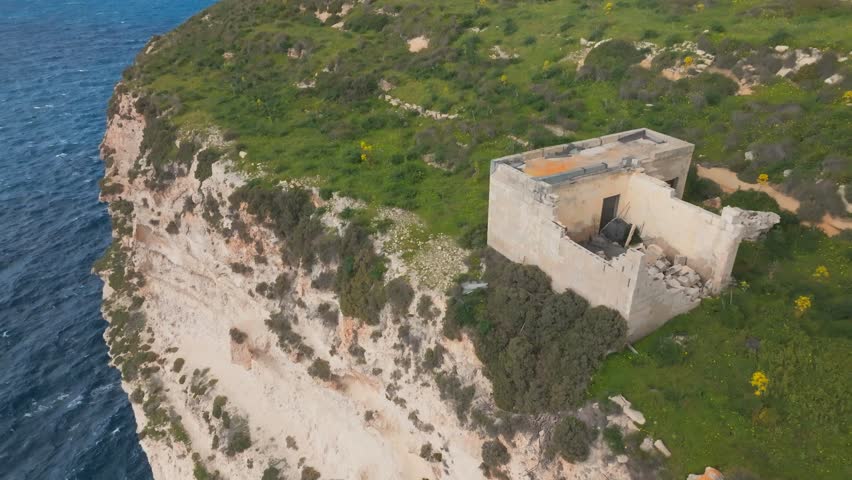 Aerial view of a rocky coastal cliff topped with dense green shrubs and yellow flowers surrounding an old, partially ruined building near the sea.