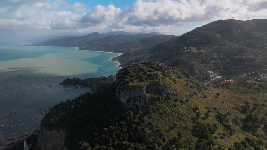 Aerial footage of a coastal town showcasing rugged cliffs, green hills, and densely packed red-roofed buildings along the shoreline under a partly cloudy sky.