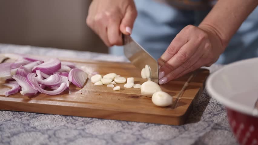 Close-up of hands slicing fresh garlic cloves and red onion on a wooden cutting board. Home cooking preparation with fresh ingredients for a rustic homemade meal.