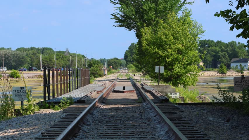 Low angle eye-level shot looking down empty railroad tracks crossing a small bridge over an algae-covered lake, with no trespassing signs and green trees on a sunny summer day.