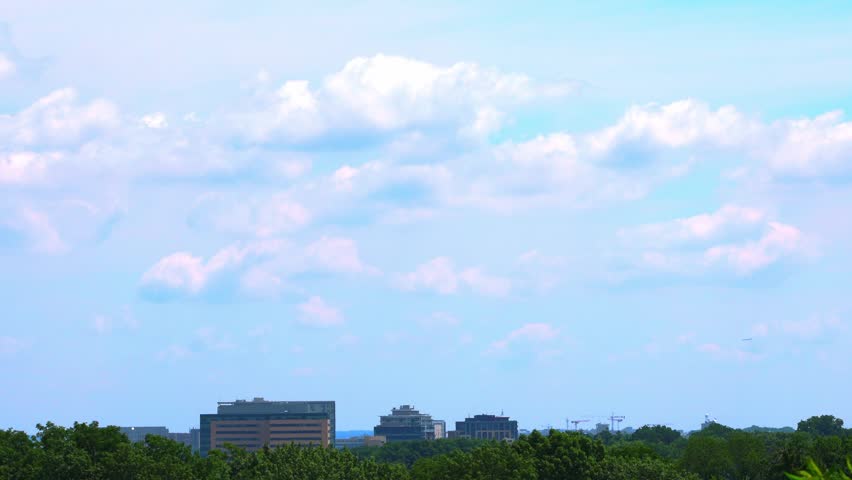 Timelapse view of the Madison, Wisconsin city skyline, featuring the UW Health University Hospital and medical campus buildings rising above a lush green tree canopy under a bright cloudy sky.