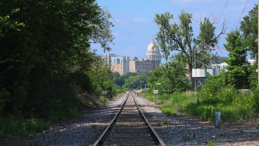 Scenic view of the Madison, Wisconsin city skyline and State Capitol dome framed by green trees and empty railroad tracks leading into the downtown area on a sunny summer day.