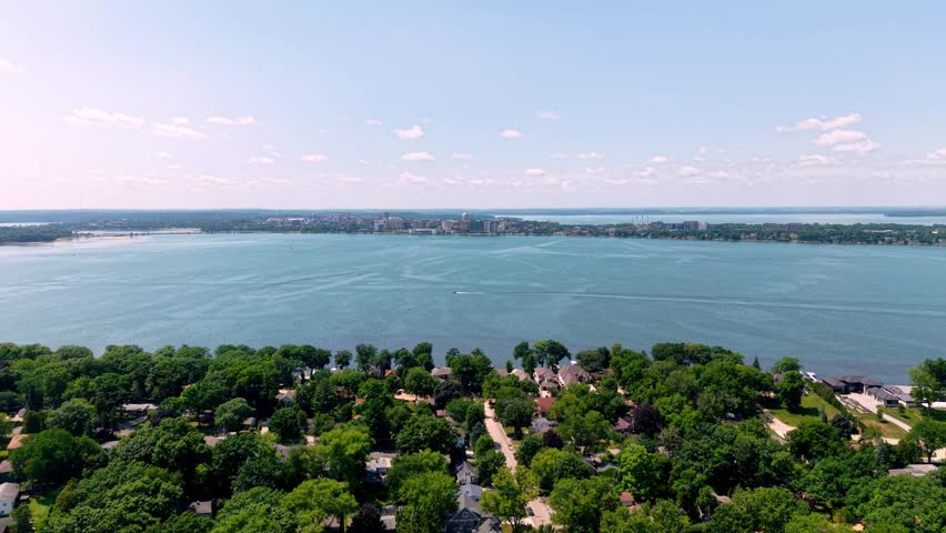 Aerial drone shot over a lush green residential neighborhood on the shore of a vast blue lake, with a boat cruising and the Madison, Wisconsin city skylinevon the distant horizon on a sunny summer day
