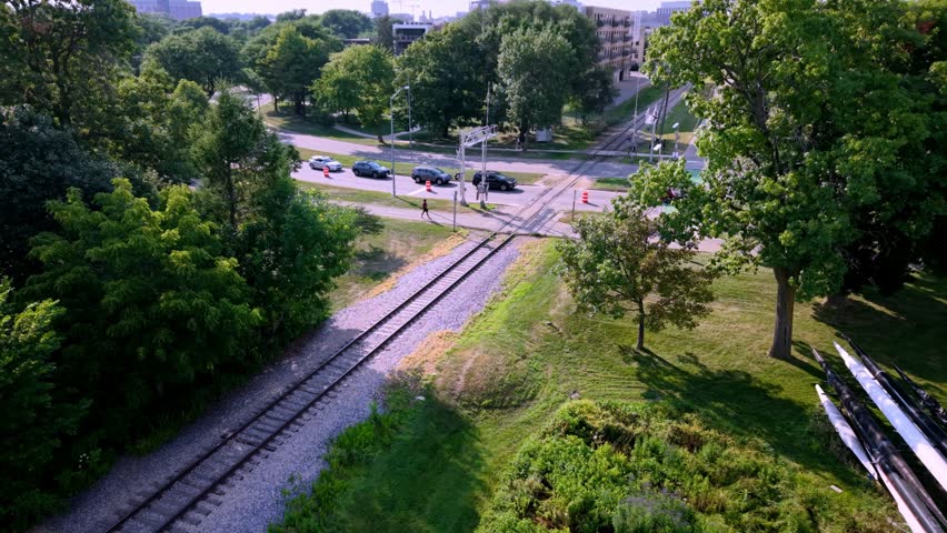 Aerial drone shot looking down at a railroad track intersecting a city street, featuring traffic, a bicycle path, pedestrians, and lush green trees on a sunny day in Madison, Wisconsin.