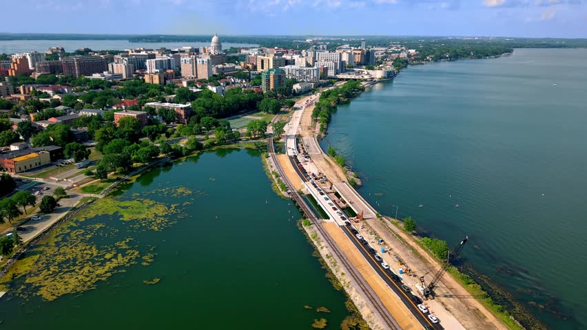 Aerial drone hyperlapse flying over a lake causeway road and railway under construction, leading towards the downtown city skyline and State Capitol building in Madison, Wisconsin on a sunny day.