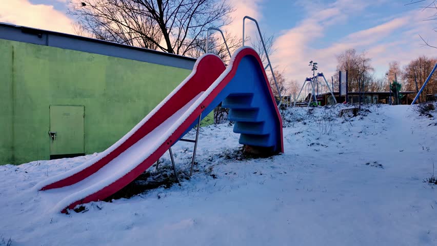 Ground-level winter photograph showing a snowy playground with a prominent red slide, blue steps, metal swings in the background, surrounded by trees and buildings under a cloudy, pink-blue sky.