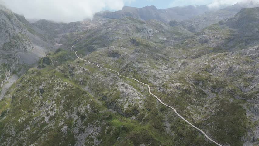 Aerial drone push in shot revealing a picturesque mountain trail snaking through the rugged, green landscape of Picos de Europa in Asturias, Spain, under a cloudy sky.