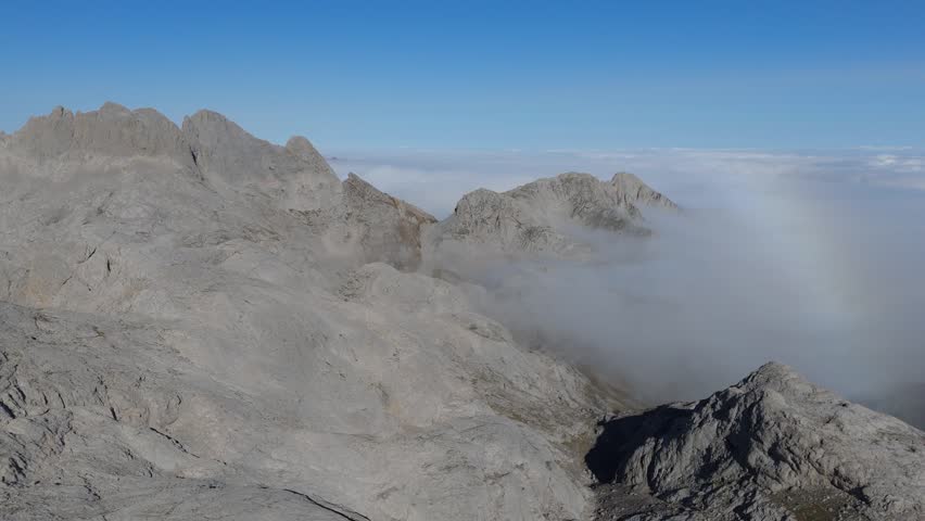 Aerial drone push in shot above the rugged Picos de Europa mountains in Asturias, Spain. Clouds drift through the valleys and around Pico de Dobresengos, revealing stunning rocky peaks.