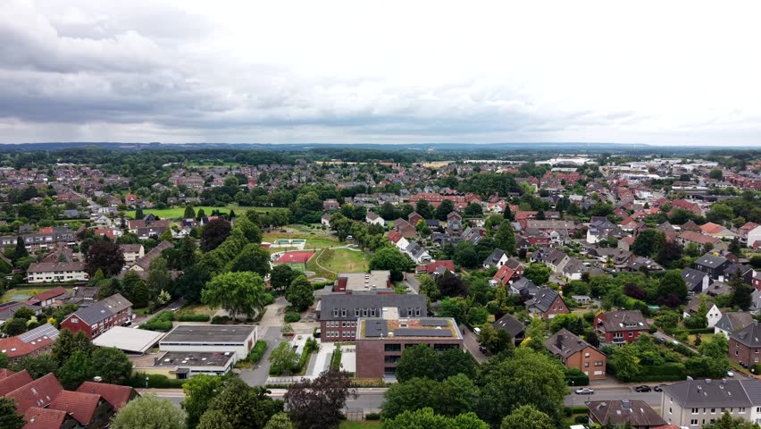 Modern school building with solar rooftop sits among dense residential rooftops in German town, sports field and tennis court stretch beneath heavy cloudy overcast sky. Aerial drone wide shot dolly.
