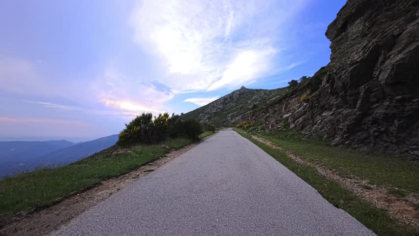 A paved mountain road curves alongside a rugged rock face beneath a sky filled with layered clouds. Grass and low shrubs line the shoulder, with a distant valley visible beyond the bend.