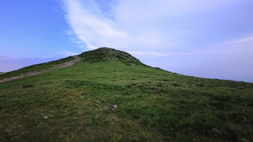 A narrow footpath crosses open grassland toward a rounded mountain summit. The trail follows the exposed ridge through a high elevation meadow beneath soft clouds.