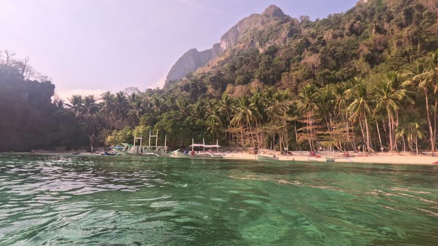 Wide shot from water of a beautiful beach surrounded by massive cliffs and lush vegetation in the Philippines. Boats and kayaks moored on the sandy shore under bright sunlight.