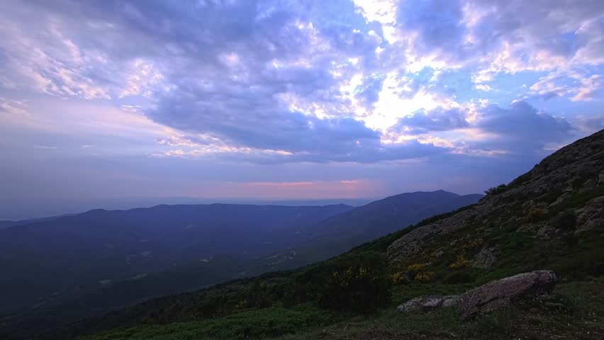 Layered clouds open above a mountain valley and rugged rocky hillside. Backlit cloud edges, distant ridges and a pink twilight horizon stretch across the evening landscape.