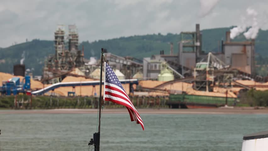 American flag waves above water near industrial pier, hazy pulp mill complex with silos plus conveyor towers lining Longview, Washington river waterfront under gray sky. Aerial drone view.