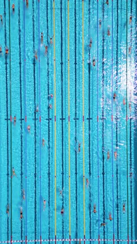 Aerial top-down view of people swimming in dedicated lanes of a large outdoor swimming pool, filled with clear blue water on a bright sunny day