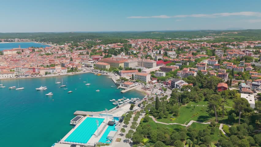 Drone turning in place over Rovinj, revealing a wide panorama of the old town and Adriatic, with a boat gliding across the water and leaving a white foam trail.