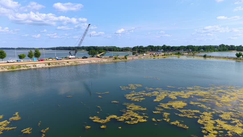 Aerial drone shot of a causeway road with active crane construction crossing a large lake, showing green algae blooms in the water on a sunny summer day in Madison, Wisconsin.