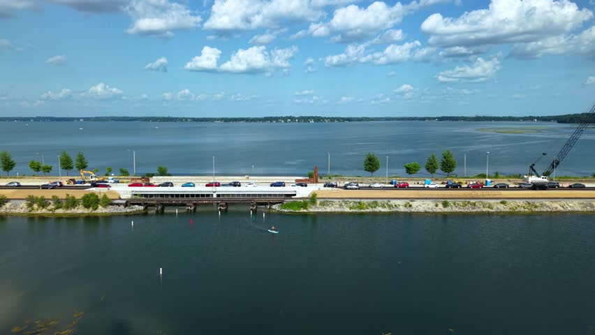 Aerial drone shot of a causeway highway bridge under construction crossing a large blue lake, with traffic moving and a person paddleboarding in Madison, Wisconsin.