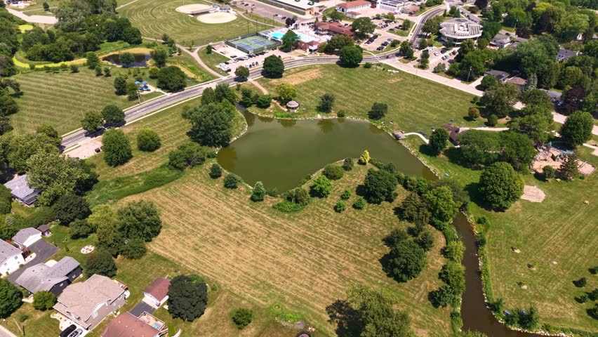 Aerial drone shot of a large suburban community park featuring a green pond, winding stream, baseball diamonds, tennis courts, and surrounding residential houses on a sunny summer day.