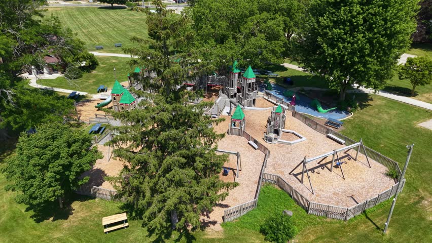Aerial drone shot of a large, empty wooden playground designed like a castle with towers, slides, and swings, surrounded by lush green trees and grass in a public park on a sunny day.