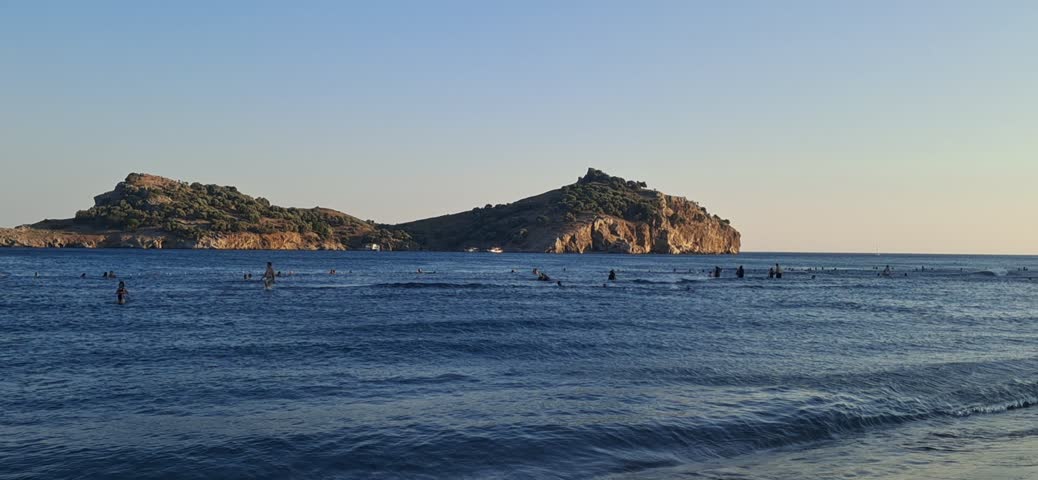 A panning shot of people swimming in the sea at Sarigerme Beach with island view in Ortaca, Mugla, Turkey during sunset.