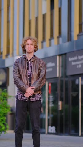 Stylish young man in a leather jacket standing outdoors in front of a modern office building, talking naturally and smiling with a light laugh. Authentic urban lifestyle scene capturing positive emotions, confidence, communication, and contemporary city living.