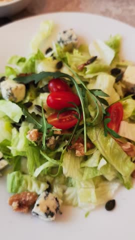 Close up view of a person eating a healthy green salad with a fork. The dish includes lettuce, arugula, cherry tomatoes, walnuts, and blue cheese