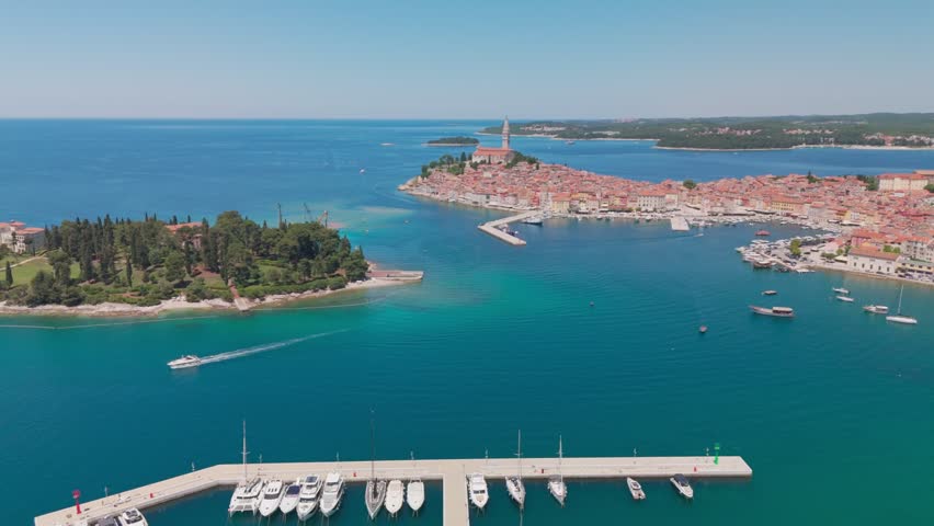 Drone moves forward after a rotation, passing a gliding boat leaving foam, revealing Katarina Island and heading toward Rovinj’s old town with the church tower above the peninsula.