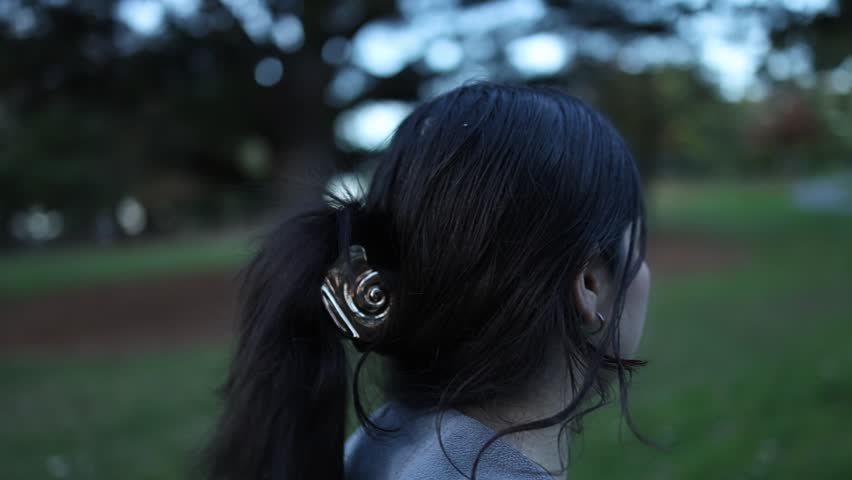 A young woman with dark hair in a metallic hair clip walks through a lush green park at dusk, her back to the camera. Soft evening light creates a serene, introspective mood.