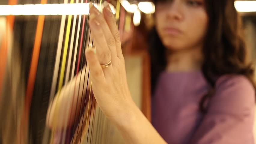 Artistic close-up shot of a woman delicately playing harp strings, her hand in focus with warm bokeh lighting. Captures elegant motion, musical concentration, and vibrant texture detail.