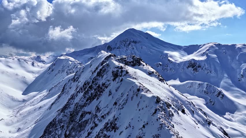 Aerial view of snow-covered mountain peaks under a bright blue sky with dramatic clouds in Nauders, Tyrol, Austria.